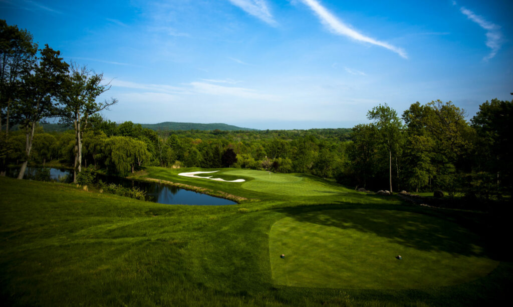 Green golf course with small pond on left, sunny day, forest and mountains at the horizon.